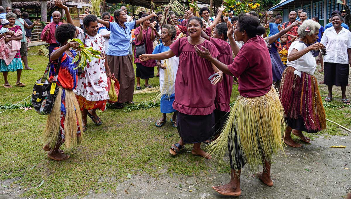 /></figure>
<p>Teachers guided their students in practicing traditional dances for the celebration, and boats ferried visitors and choir groups in from nearby islands. Every detail, from the replica of a traditional house that would carry the New Testament to the vibrant dance routines, was planned with love and anticipation.</p>
<h3>The Big Day Arrives</h3>
<p>The day of the dedication began bright and sunny. Groups of villagers arrived on foot or boat, all of them carrying food — pigs and garden produce — for the day’s feast. They were escorted to the celebration grounds next to the church with dancing and joyful shouts. While a downpour delayed the start of the celebration by a few hours, at 11 a.m., the festivities officially began!</p>
<p>Four Saliba translators kicked off a grand procession, carrying in the replica house that contained the New Testament. Dancers in traditional attire led the way.</p>
<figure class= The Saliba community celebrating the New Testament through song and dance.